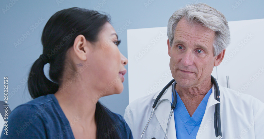 Wall mural Close up of senior Caucasian male doctor listening to Asian female patient in exam room. Older man medical professional and Japanese woman talking in hospital