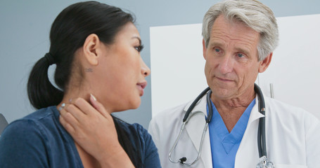 Close up of Senior Caucasian male doctor asking female patient about neck pain in exam room. Medical professional listens to Asian woman explain spinal trouble in hospital