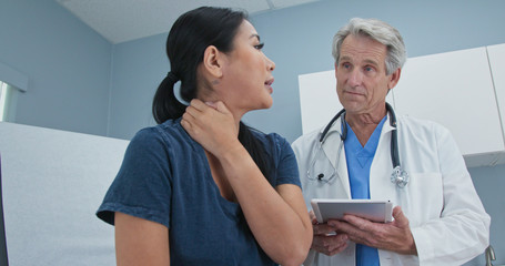 Low angle side view of Asian woman explaining neck pain to doctor in exam room. Senior Caucasian male medical professional listening to patient with spinal problem