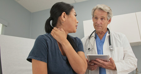 Obraz premium Low angle side view of Japanese woman explaining neck pain to doctor in exam room. Senior Caucasian male medical professional listening to patient with spinal problem
