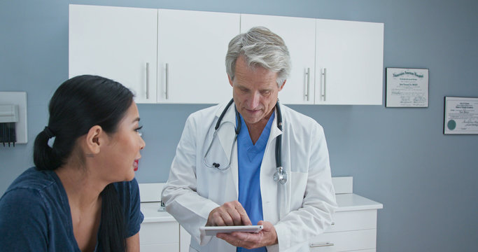 Older Man Doctor Taking Medical History On Tablet Computer From Japanese Woman In Exam Room. Senior Caucasian Male Medical Professional In OBGYN Office With Patient