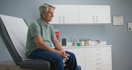 Senior Caucasian male patient waiting patiently for doctor while sitting on exam room table. Older...