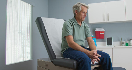 Senior Caucasian male patient waiting patiently for doctor while sitting on exam room table. Older man going to regular appointment for annual check up