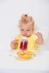 Cute little baby girl 1 year old eating cereal flakes and drinking juice or compote from a bottle at the table isolated on white background