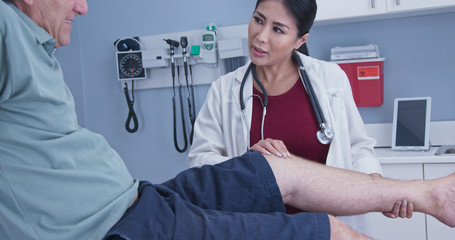 Senior man with hurt knee getting it checked by female medical professional. Physical therapist or doctor with older male patient in exam room checking range of motion on knee injury