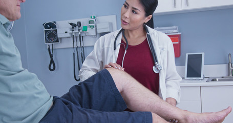 Patient with hurt knee getting it checked by female medical professional. Physical therapist or doctor with older male patient in exam room checking range of motion on knee injury