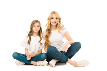 smiling mother and daughter sitting on floor with crossed legs and looking at camera on white background