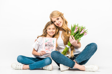 mother with bouquet of pink tulips and daughter with happy mothers day greeting card sitting on floor with crossed legs on white background