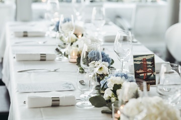 Wedding table decoration. Floral garland of greenery and blue flowers lies between glasses on the white table