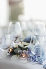 Fototapeta na wymiar Wedding table decoration. Floral garland of greenery and blue flowers lies between glasses on the white table
