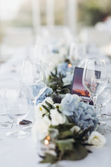 Wedding table decoration. Floral garland of greenery and blue flowers lies between glasses on the white table