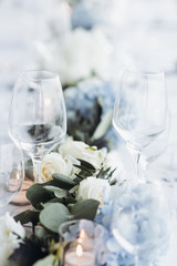Wedding table decoration. Floral garland of greenery and blue flowers lies between glasses on the white table