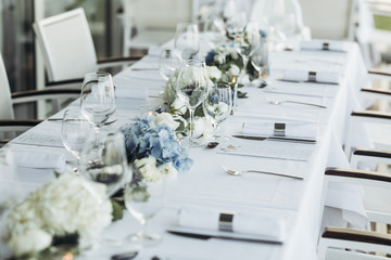 Wedding table decoration. Floral garland of greenery and blue flowers lies between glasses on the white table
