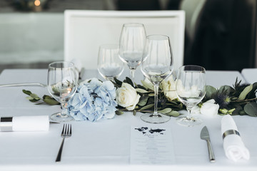 Wedding table decoration. Floral garland of greenery and blue flowers lies between glasses on the white table