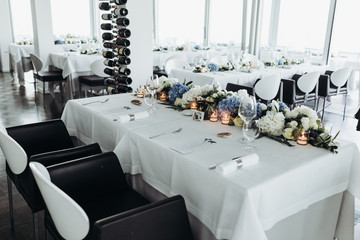 Wedding table decoration. Floral garland of greenery and blue flowers lies between glasses on the white table