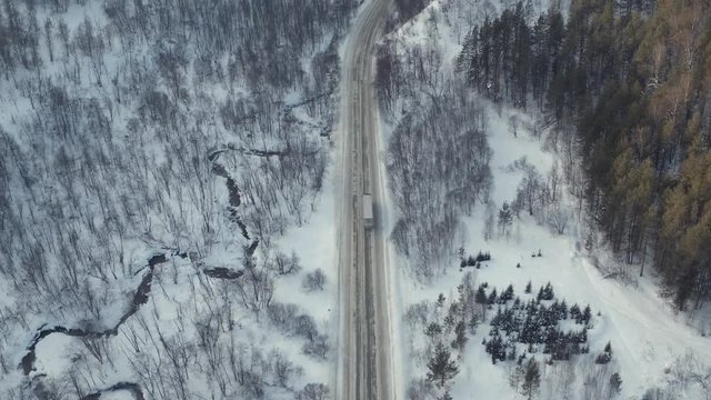 Alone truck with white clear empty open trailer is driving along a winding frost road in the coniferous forest, mountains in winter frozen sunny day - Aerial Drone View