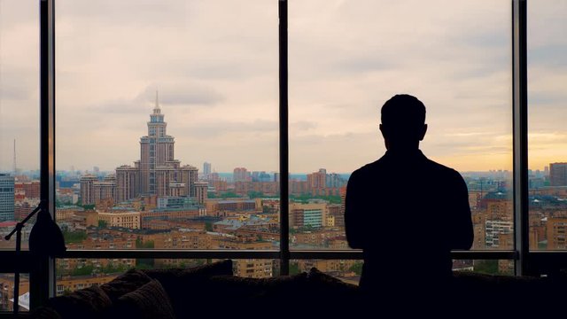 silhouette of man wearing polo shirt enters frame, slowly confidently coming to window with beautiful view of Moscow from his apartment office. thoughtfully looking at cloudy city. Medium slow motion 