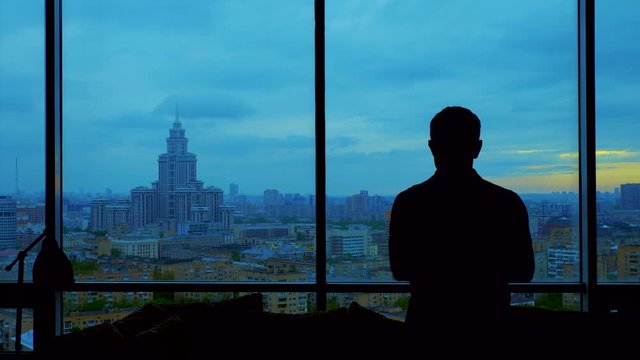 silhouette of man wearing polo shirt enters frame, slowly confidently coming to window with beautiful view of Moscow from his apartment office. thoughtfully looking at cloudy city. Medium slow motion 
