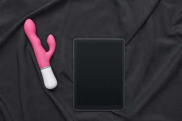 Shopping online. Top view of pink vibrator and digital tablet against of black silk fabric.