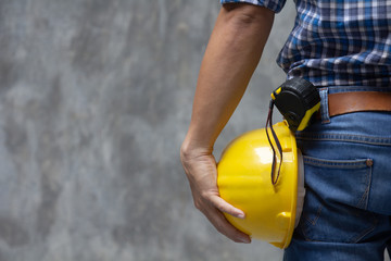 Male Architect worker holding yellow safety helmet in hand outdoor over concrete wall background, Close up back view with copy space. engineer man in casual wearing jeans at construction site factory.