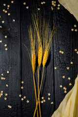 Horizontal composition still Life with wheat ears on a background of black burnt boards with scattered pastry stars and beige fabric. The concept of family happiness