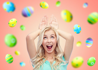 easter, holidays and people concept - happy smiling young woman making bunny ears over pink or living coral background with colored eggs