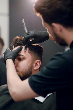 Young brunette man is getting shaved with a razor by a hairdresser at the barbershop.