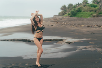 Slim beautiful girl in swimsuit posing with raised arms on beach with black sand. Traveler resting on vacation at ocean