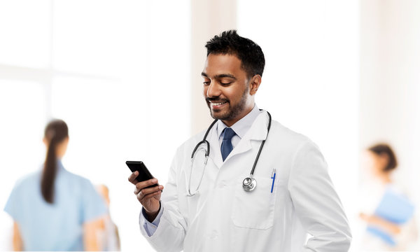 medicine, technology and healthcare concept - smiling indian male doctor in white coat with smartphone and stethoscope over hospital background