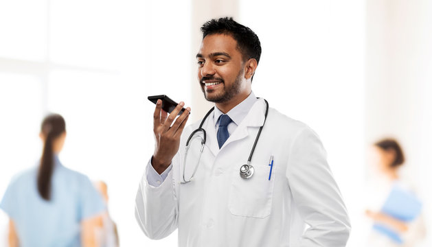 medicine, technology and healthcare concept - smiling indian male doctor in white coat with stethoscope using voice command recorder over hospital background
