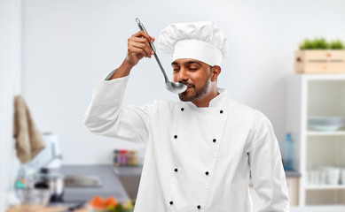 cooking, profession and people concept - happy male indian chef in toque tasting food from ladle over kitchen background