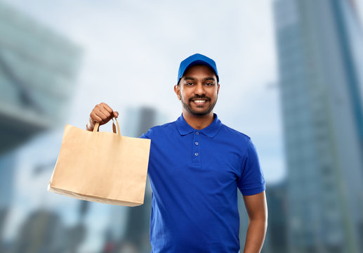 Service And People Concept - Happy Indian Delivery Man Food In Paper Bag In Blue Uniform Over Tokyo City Skyscrapers Background