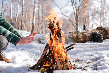 Basking near a campfire in a snowy birch forest. Female person near a fire on a sunny winter day in the woods