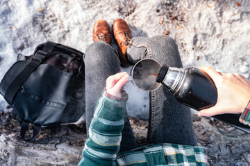 Pouring hot drink out of thermos at a campsite. Person in a winter forest during a hiking trip...