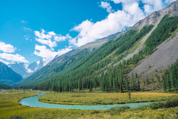 Serpentine river in valley before beautiful glacier. Snowy rocks behind mountains with conifer forest. Huge clouds on giant snowy mountain top under blue sky. Atmospheric landscape of highland nature.