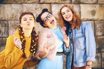 Three beautiful girls models eat lollipops. Girlfriends with different hairstyles and styles. Girls send kisses, wriggles, laugh, have fun. Girls in sunglasses and fashionable clothes outside. Cute 