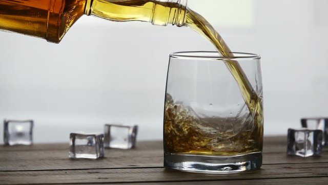 Whiskey pouring into a glass with ice cubes on old wooden table in slow motion. Seamless cinemagraph video