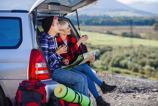 Young friends travelers sitting on trunk of car with cap of tea and looking at the paper map. Two happy hipster girls enjoying on vacation in mountains. Summer travel concept.