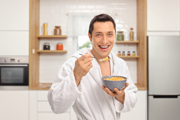 Smiling young man in a dressing gown eating cereal from a bowl in a kitchen