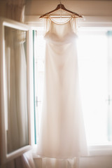 Beautiful wedding dress hangs before the bright window