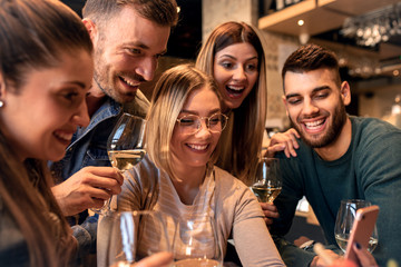 Group of young friends having fun in restaurant, they are looking at smartphone and making selfie.