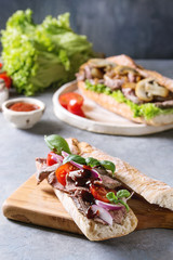 Beef baguette sandwich with tomatoes, basil, red onion served on ceramic plate with ingredients above over grey blue table.