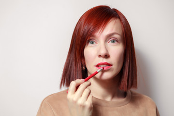 Closeup portrait redhead pretty girl paints her lips with red pencil lipstick. Concept school of makeup, future makeup artist, get ready for party