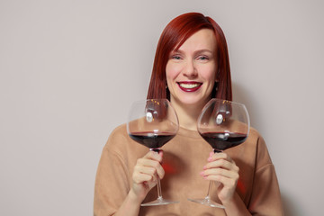 Young cheerful redhaired girl hold and get fun with two glasses of red wine. Concept alcoholism, alcohol addiction, bad habit