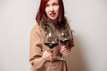 Young funny redhaired girl sommelier with fur cape on white background smile, hold two glasses red wine. Concept alcoholism, addiction, bad habit, collection great rare wines, winelover, expert