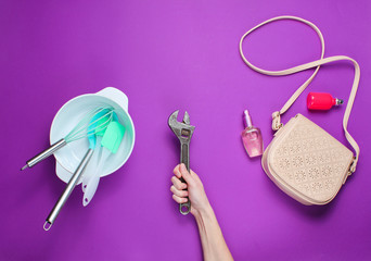 Women choose male work. Female hand holding a wrench on the background of bowl with kitchen tools, bags and bottle of perfume. Purple background, top view