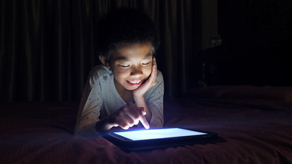 Asian boy lying in bed at night and playing digital tablet.