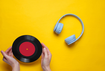 Women's hands hold vinyl record on yellow background with over-ear headphone. Pop culture, retro style, 80s. Minimalism. Top view. Flat lay