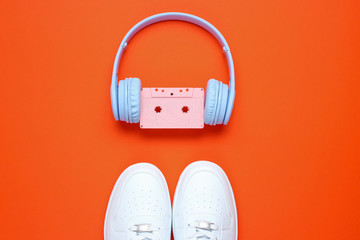 Retro style. 80s. Pop culture. Minimalismalism. Headphones with audio cassette, white sneakers on   orange background. Top view