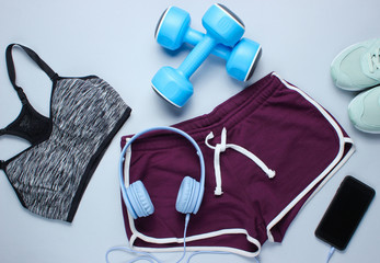 Sportswear and fitness equipment on gray background. Smartphone with headphones, plastic dumbbells, sports bra, shorts, sneakers on gray background. Top view
